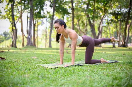 Beautiful Asian woman in sportswear doing One legged plank on the knee pose, practicing yoga and cardio on her weekend at the park.