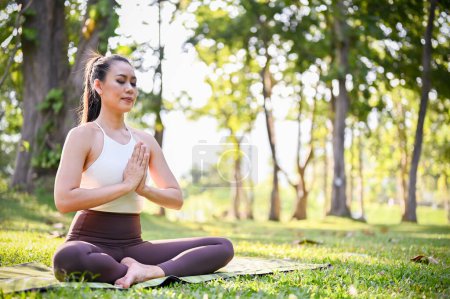 Téléchargez les photos : Belle et charmante femme asiatique en tenue de sport assise sur son tapis de yoga dans le jardin, se concentrant sur son souffle, méditant avec pose de phoque de salutation. - en image libre de droit