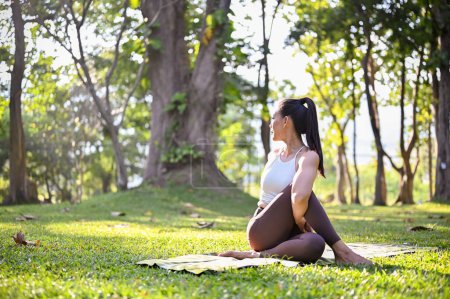 Téléchargez les photos : Femme asiatique attirante et sportive en vêtements de sport faisant demi-seigneur de la pose pêchée, pratiquant le yoga dans le beau parc verdoyant le matin. Concept de style de vie bien-être - en image libre de droit