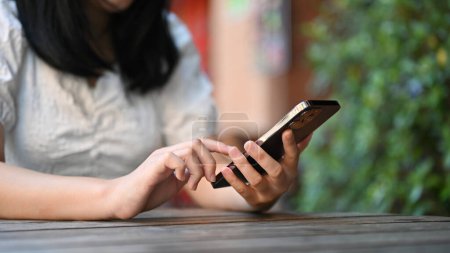 disparo recortado, Joven mujer asiática usando su teléfono inteligente mientras está sentado en la cafetería al aire libre.