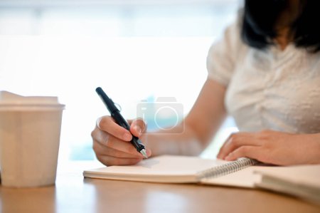 Téléchargez les photos : Jeune étudiante asiatique ou pigiste écrivant son journal ou prenant des notes sur son carnet, travaillant à distance au café. image recadrée et en gros plan - en image libre de droit