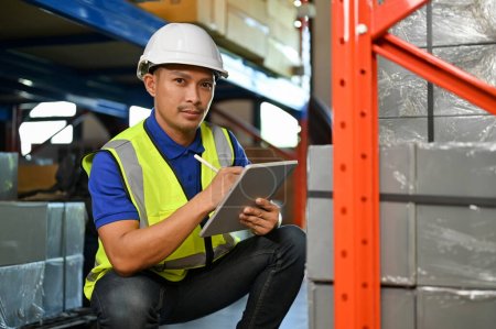 Photo for Handsome and professional Asian male warehouse worker or foreman in white hardhat and uniform, checking an inventory list on tablet, working in warehouse. shipping, cargo, factory concept - Royalty Free Image