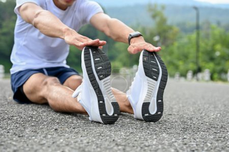 Téléchargez les photos : Homme sportif asiatique actif et beau en vêtements de sport et chaussures de course étirant les jambes, se réchauffant avant de courir en plein air. image recadrée - en image libre de droit