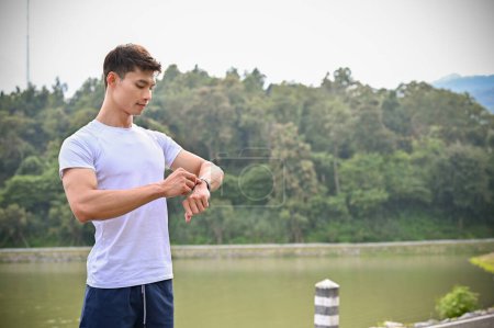 Téléchargez les photos : Beau et en forme jeune homme athlétique asiatique en vêtements de sport, vérifier ses miles de course et brûler des calories sur sa smartwatch. Concept gadget sport - en image libre de droit
