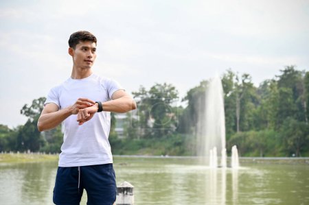 Téléchargez les photos : Beau jeune homme athlétique asiatique en vêtements de sport avec sa montre intelligente, debout à l'extérieur, le comptage des calories, se préparer à courir. Activité saine avec concept technologique - en image libre de droit