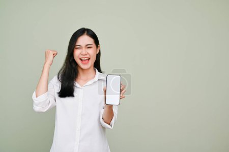 Photo for Happy and charming young Asian woman in white shirt showing a smartphone white screen mockup and fist up, standing against grey background. - Royalty Free Image