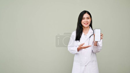 Téléchargez les photos : Charmant médecin asiatique femme en robe blanche paume ouverte montrant une maquette d'écran blanc smartphone pour ajouter votre bannière graphique, debout sur fond gris. recommander une nouvelle application, promotion chaude - en image libre de droit