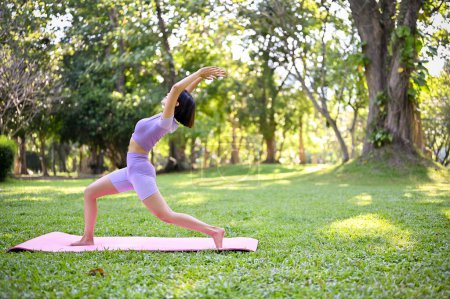 Fit and active young Asian woman in sportswear stretching her legs, practicing yoga in warrior I pose in the park. Wellbeing and healthy lifestyle concept