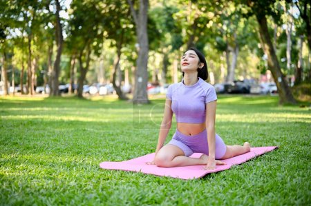 Téléchargez les photos : Belle jeune femme asiatique en tenue de sport pratiquant la pose de yoga demi-pigeon sur son tapis de yoga dans le parc vert. Concept d'exercice de yoga - en image libre de droit