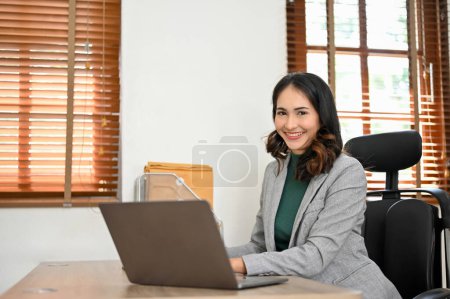 Photo for Attractive millennial Asian businesswoman in formal suit smiling and looking at camera while sitting at her desk in modern office. - Royalty Free Image
