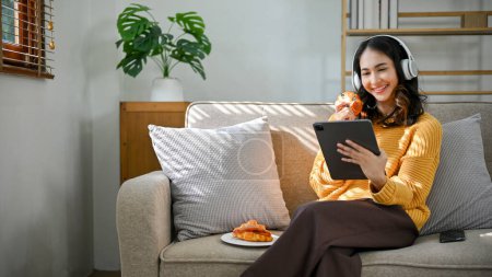 Téléchargez les photos : Attrayant jeune asiatique femelle en pull jaune portant des écouteurs, regarder quelque chose sur tablette et manger des boulangeries délicieuses tout en se relaxant dans son salon. - en image libre de droit