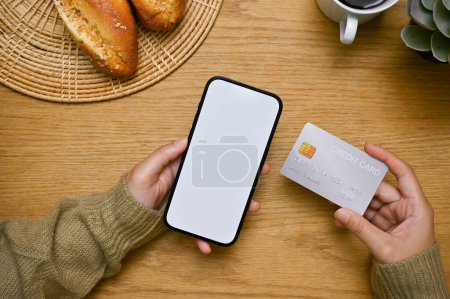Photo for A female hands holding a smartphone white screen mockup and a credit card over wooden tabletop. top view - Royalty Free Image