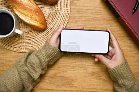 Photo for A smartphone white screen mockup in a horizontal position is in a woman's hand over wooden tabletop. top view, watching video, playing mobile game or using mobile application - Royalty Free Image
