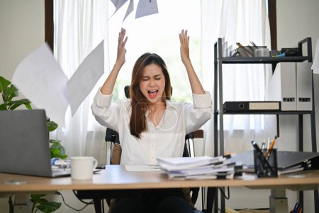 Enfurecida, furiosa, loca y loca millennial empresaria asiática o trabajadora de oficina gritando, gritando y vomitando papeles en su escritorio de oficina.