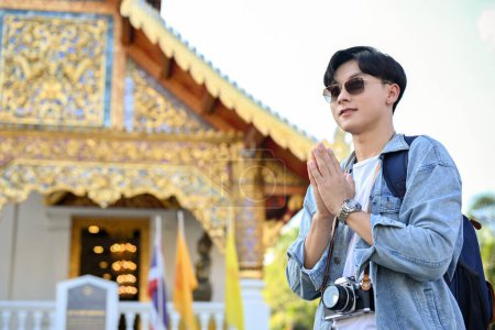 Photo for Handsome young Asian male traveler wearing sunglasses, standing and putting his hands together in a prayer position over blurred background of a beautiful Thai's temple. Vacation concept - Royalty Free Image