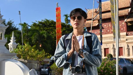 Photo for Young Asian male traveler wearing sunglasses, standing and putting his hands together in a prayer position over blurred background of a beautiful Thai's temple. Asia traveling concept - Royalty Free Image