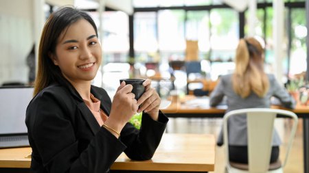 Téléchargez les photos : Magnifique et professionnelle jeune femme d'affaires asiatique en costume noir formel se trouve au café, tenant une tasse de café et souriant à la caméra. - en image libre de droit