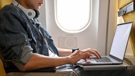 Téléchargez les photos : Un jeune voyageur asiatique branché utilisant son ordinateur portable, travaillant sur sa tâche pendant son voyage dans l'avion. image recadrée - en image libre de droit