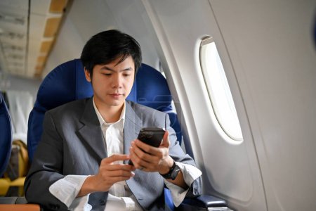Téléchargez les photos : Jeune homme d'affaires asiatique intelligent et séduisant utilisant son smartphone pendant le vol en jet privé pour son voyage d'affaires. - en image libre de droit