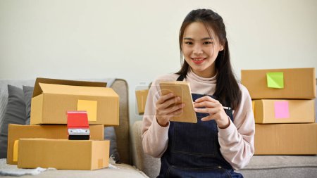 Foto de Bonita y encantadora joven asiática propietaria de una tienda en línea se sienta en su sala de estar, revisando sus cajas de envío, preparando sus pedidos de envío. - Imagen libre de derechos