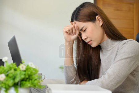 Stressed and upset young Asian woman sits at her working desk, feeling tired about her work, suffering from a headache or migraine, failing at her work, getting a job complaint email.
