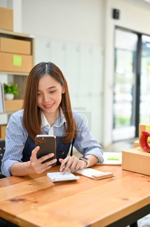 Téléchargez les photos : Portrait, belle et charmante jeune femme asiatique e-commerce démarrage d'entreprise se trouve dans son bureau, en utilisant son smartphone pour vérifier ses commandes en ligne. - en image libre de droit