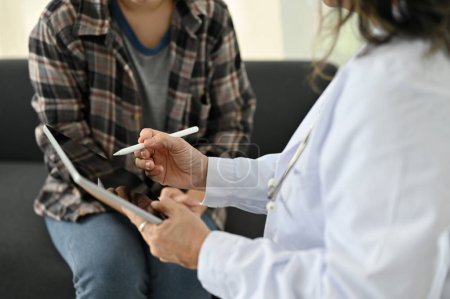 zugeschnittenes und nahes Bild, professionelle asiatische Ärztin mit digitalem Tablet, um die Symptome der Patientin aufzuzeichnen, mit einem Gespräch mit ihrem Patienten in der Klinik.