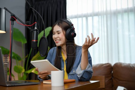 Photo for Cheerful and happy young Asian female radio host or online influencer is recording and board-casting her online radio channel in her home studio. freelance content creator concept. - Royalty Free Image