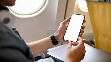 Foto de Un pasajero viajero masculino se sienta en el asiento de la ventana en clase económica, usando su teléfono inteligente, sosteniendo una maqueta de pantalla blanca del teléfono móvil. imagen de primer plano - Imagen libre de derechos