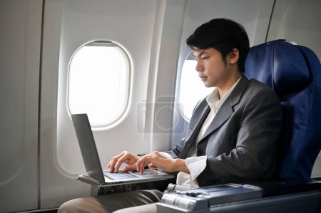 Téléchargez les photos : Homme d'affaires asiatique professionnel travaillant, gérant ses tâches professionnelles sur ordinateur portable pendant le vol pour son voyage d'affaires. Concept d'entreprise et de transport - en image libre de droit