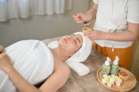 Photo for Relaxed and peaceful young Asian woman lying on massage table with eyes closed, getting facial treatment with special facial detoxifying clay mask. - Royalty Free Image