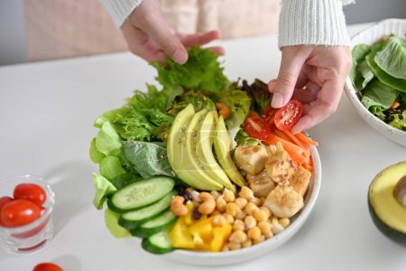 Foto de A female making a plate of Buddha bowl or plant based salad vegetables mixed, grilled tofu with chickpea, beans, tomatoes, avocado slides, cucumber, and green salad. diet food concept - Imagen libre de derechos