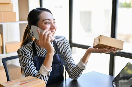 Photo for Charming and cheerful millennial Asian female online shop owner or entrepreneur talking on the phone with her suppliers while working in her packing room. - Royalty Free Image