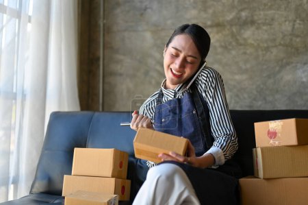 Foto de Charming and busy millennial Asian female e-commerce business entrepreneur or online seller is on the phone with her suppliers while preparing shipping packages in her living room. - Imagen libre de derechos