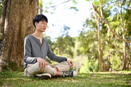 Photo for Handsome and relaxed young Asian man in casual clothes meditating under the tree in the greenery park. - Royalty Free Image
