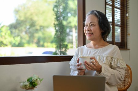 Photo for Happy aged Asian businesswoman working from home, looking out the window and daydreaming - Royalty Free Image