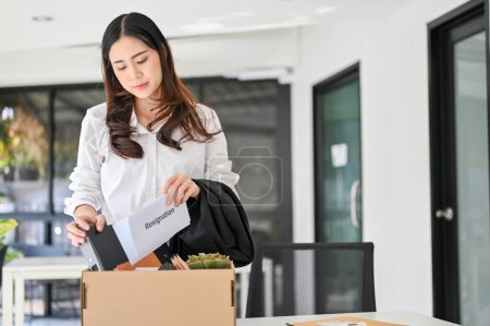 Foto de Sad and depressed young Asian female office worker packing her stuff into a cardboard box in the office, quitting her job or getting fired from her boss. unemployment, resign, career failure - Imagen libre de derechos