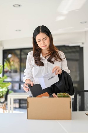 Foto de Sad and crying young Asian female office worker packing her stuff into a cardboard box in the office, being fired from her boss. unemployment, resign, career failure - Imagen libre de derechos