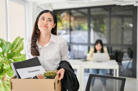 Foto de Happy and fearless Asian woman carrying her personal stuff and resignation letter in a cardboard box, excited to resign from an unprofessional company. happy fired woman, transition to a better job - Imagen libre de derechos