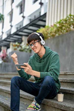 Photo for Portrait, Cheerful young Asian male college student in casual clothes wearing headphones and using his smartphone on street stairs, watching football match. - Royalty Free Image