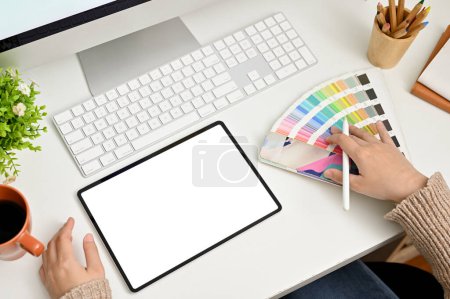Foto de Vista superior de una diseñadora gráfica femenina que trabaja en su escritorio, eligiendo el color de su plantilla de producto de la paleta de colores, utilizando una tableta digital. maqueta de pantalla blanca del comprimido - Imagen libre de derechos