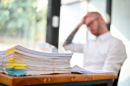 Foto de Pile of unfinished document paperwork or a stack of financial report on the table over blurred background of stressed businessman sits at the table. close-up image - Imagen libre de derechos