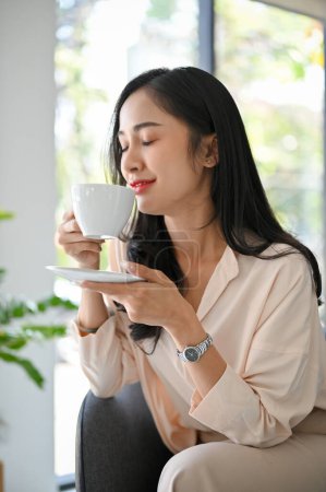 Téléchargez les photos : Attrayant et détendu millénaire asiatique femme d'affaires ou femme patron les yeux fermés, sirotant le café du matin à son bureau. - en image libre de droit