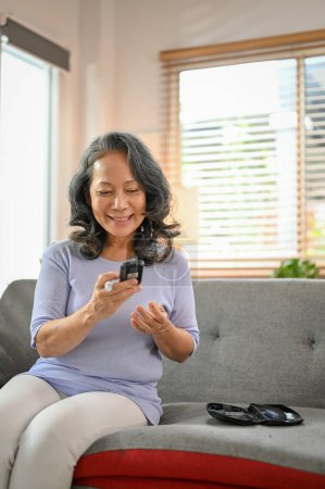 Photo for A happy 60-year-old retired Asian woman using a glucose meter to measure her blood sugar level at home. Diabetes and health care concept - Royalty Free Image