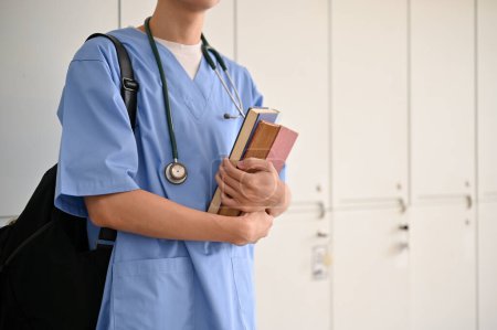 Téléchargez les photos : Cropped image, Un étudiant asiatique en médecine dans un uniforme avec ses livres médicaux et son sac à dos, se tient dans un vestiaire. - en image libre de droit