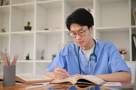 Téléchargez les photos : Smart jeune asiatique mâle étudiant en médecine dans un uniforme se concentre sur la lecture d'un livre dans la bibliothèque de l'école de médecine. - en image libre de droit