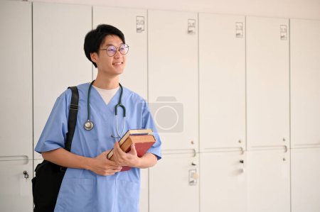 Photo for Smart and inspired young Asian male medical student in a uniform with his medical books and backpack, stands in a locker room. - Royalty Free Image