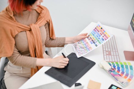Téléchargez les photos : Image en gros plan d'une jeune graphiste utilisant une tablette graphique pour esquisser un nouveau design tout en choisissant la couleur à partir d'un échantillon de nuancier. - en image libre de droit