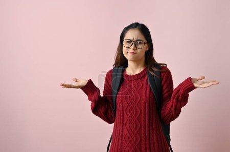 Photo for Confused and doubtful young Asian female college student shrugging her shoulders and open palms while standing against an isolated pink background. unsure, uncertain, maybe, have no idea - Royalty Free Image