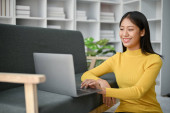 Young happy Asian woman using laptop computer while sitting on the floor and lean against sofa in the living room.  Longsleeve T-shirt #653127354